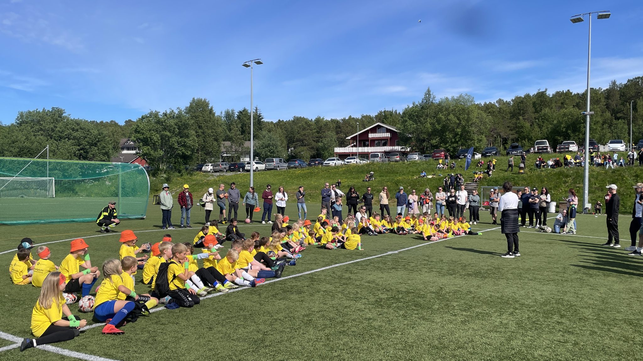 Barn i gule drakter sitter i en bue på kunstgressbanen, med foreldre og trenere bak